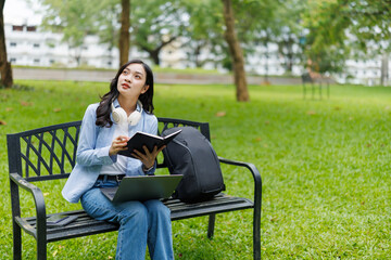 Obraz premium Young asian student is sitting on a bench in a park, taking notes on her notebook while thinking and looking up, with her laptop and backpack next to her