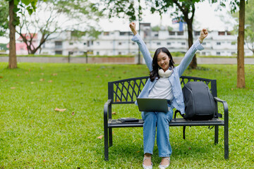 Fototapeta premium Young asian woman sitting on bench with laptop raising arms in excitement celebrating success in distance learning, freelance work or scholarship achievement in green city park