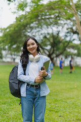 Portrait of a smiling asian female student holding tablet and laptop while standing in university park with her backpack and headphones, enjoying campus life and educational opportunities