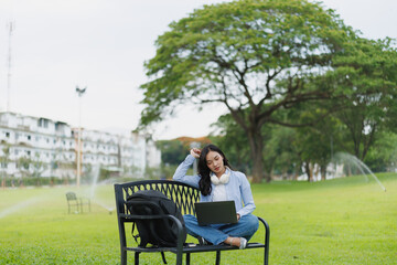 Fototapeta premium Concentrated young female student is working on laptop while sitting on bench in park with backpack near her, enjoying good weather and fresh air