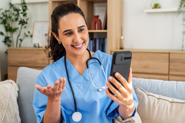 Nurse in scrubs or doctor is enjoying a friendly video call on her mobile phone with a patient or friend while sitting comfortably on a sofa. Her cheerful demeanor adds warmth to the cozy living room