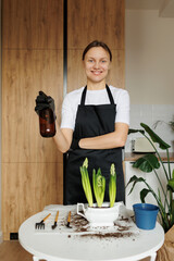 Smiling young florist woman in apron holds hyacinth flowers in new ceramic pot and waters houseplant from water sprayer at home kitchen after repotting. Seasonal work with transplantation plants.