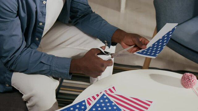 Midsection view of African American man dressed in white pants and blue shirt using scissors to cut out triangle US national flags while sitting at round table in living room