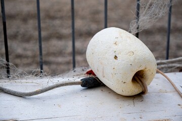 Close-up of a white buoy secured with a rope, resting on an outdoor surface next to a fishing net. The scene suggests a nautical environment with a focus on marine equipment.
