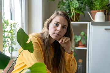 Portrait of thoughtful middle aged woman surrounded by indoor plants, looking at camera. Tired serious blonde female plant lover with green thumb, home gardening passion, botanical enthusiast. 