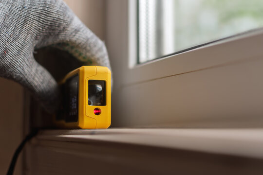 A gloved hand holds a yellow laser distance meter against a window sill, focusing on precise measurement in home renovation or construction projects.