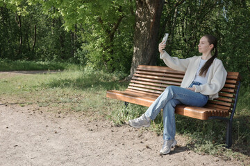 Obraz premium Young woman sitting on a bench in a sunny park, taking selfie with smartphone. Concept of digital communication, leisure, outdoor lifestyle, springtime relaxation and solo travel.