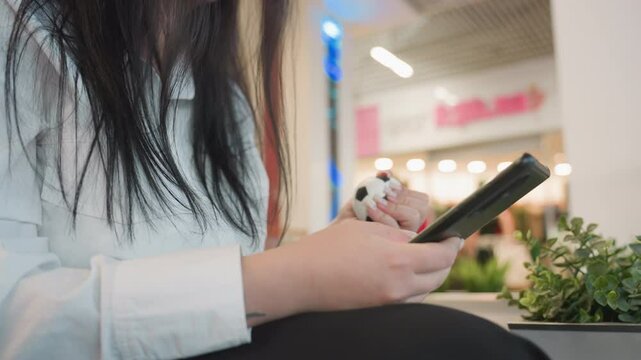 Young woman with headset seated indoors operating digital device while gently squeezing stress ball, wearing glasses with long dark hair, blurred storefront in background