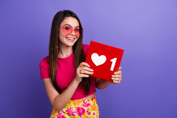 Smiling young woman holding a social media love symbol against a vivid purple background,...