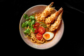 A bowl of ramen with tempura shrimp, soft-boiled egg, cherry tomatoes, cilantro, and noodles in a rich broth. The vibrant colors and textures make it visually appealing and appetizing