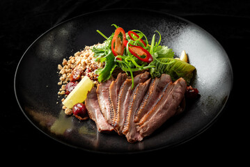 Sliced duck breast with fresh herbs, red chili, crushed nuts, and lemon wedge on a black plate. Gourmet dish with elegant presentation, highlighting rich flavors and textures.