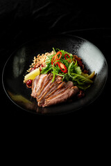 Sliced duck breast with fresh herbs, red chili, crushed nuts, and lemon wedge on a black plate. Gourmet dish with elegant presentation, highlighting rich flavors and textures.