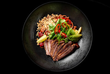 Sliced duck breast with fresh herbs, red chili, crushed nuts, and lemon wedge on a black plate. Gourmet dish with elegant presentation, highlighting rich flavors and textures.
