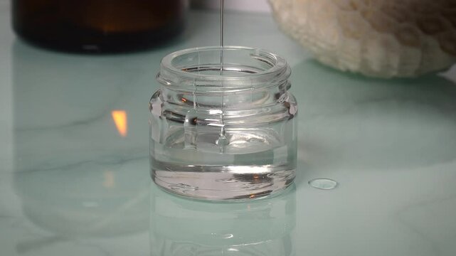 Transparent gel-like liquid being poured into a glass jar against a blue glossy background. Skincare or cosmetic product preparation. Clean, modern, and minimalistic beauty concept.