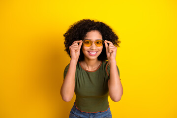 Joyful young woman in stylish outfit with curly hair wearing yellow sunglasses against vibrant...