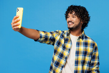 Young happy Indian man wear yellow checkered shirt casual clothes doing selfie shot on mobile cell phone post photo on social network isolated on plain blue cyan background studio. Lifestyle concept.