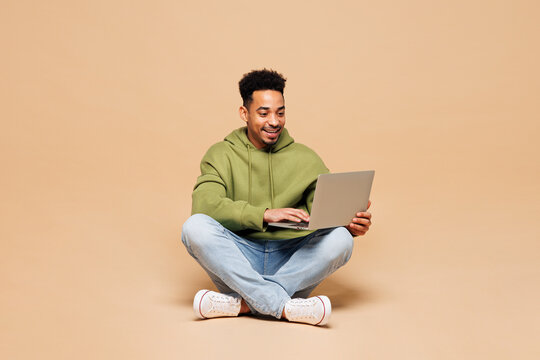 Full body young smart IT man of African American ethnicity wears green hoodie casual clothes sit hold use work on laptop pc computer isolated on plain pastel light beige background. Lifestyle concept.