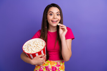 Young woman enjoying popcorn with an enthusiastic expression on purple background, wearing vibrant...