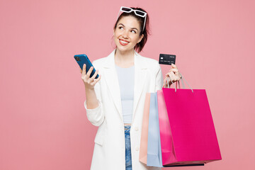 Young woman wear white clothes hold paper package bags after shopping, credit bank card order delivery use mobile cell phone isolated on plain pink color background. Black Friday sale buy day concept.