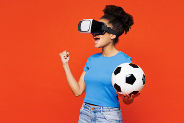 Young winner woman fan wears blue t-shirt cheer up support football sport team hold in hand soccer ball watch tv live stream watching in vr headset pc gadget isolated on plain red orange background.
