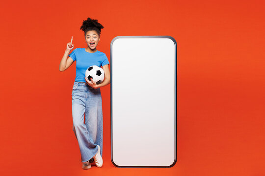 Fototapeta Full body young woman fan in blue clothes support football sport team hold soccer ball watch tv live stream big blank screen mobile cell phone point finger up isolated on plain red orange background
