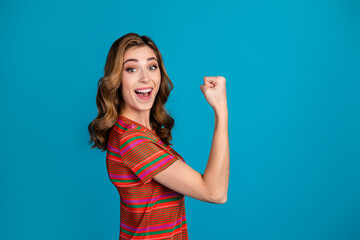 Excited young woman in colorful striped t-shirt making a victory gesture against blue background,...
