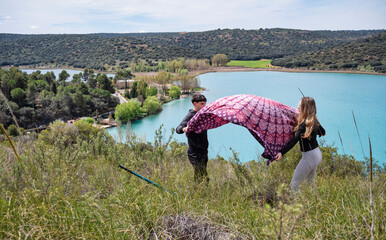 Teen girl and boy spreading a blanket on a grassy hillside overlooking a turquoise lagoon, preparing to rest and enjoy the view after a scenic outdoor hike