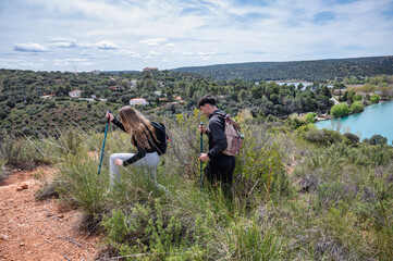 Teen girl and boy hiking through rugged hillside terrain, using walking poles to navigate a natural trail with a turquoise lagoon and village in the background