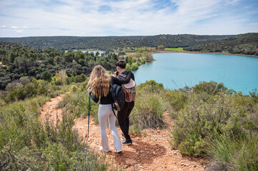 Teen girl and boy hiking along a scenic path beside a turquoise lagoon, as the boy uses binoculars to admire the landscape while the girl supports him during their nature walk