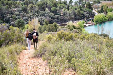 Teen boy and girl walking on a nature trail with backpacks, embracing as they hike toward a forest and turquoise lagoon on a peaceful sunny day
