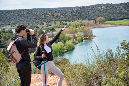 Teen boy and girl hiking with backpacks by a turquoise lagoon, using binoculars and pointing at the scenic landscape on a sunny outdoor adventure
