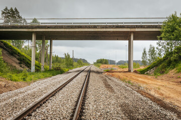 Obraz premium Symmetrical view of railway tracks stretching into the distance and passing beneath a concrete bridge, surrounded by forested terrain and cloudy skies.
