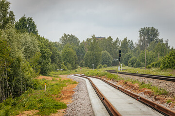 Obraz premium A curved section of newly laid railway track diverges beside an older line, with a circular signal sign and red light signal post in a green, forested area.