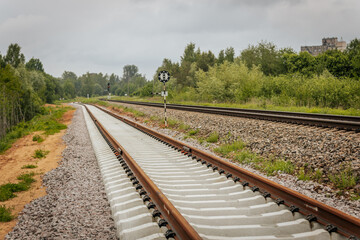 Obraz premium A curved section of newly laid railway track diverges beside an older line, with a circular signal sign and red light signal post in a green, forested area.