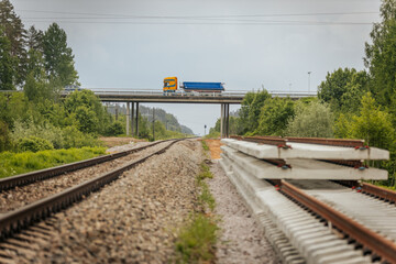 Stacked railway track sections beside operational rails with a yellow-blue truck crossing an...