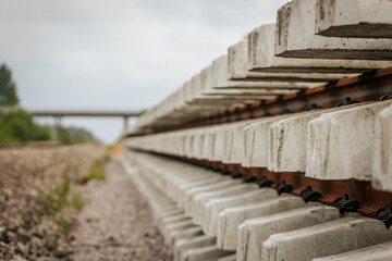 A close side view of stacked concrete railway track sections alongside a railway line, with a bridge visible in the background under cloudy skies.