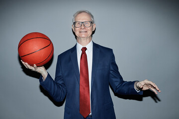 Portrait of senior Caucasian man wearing suit and tie smiling at camera while holding basketball against plain grey background in studio, camera flash