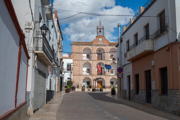 A picturesque street in Laujar de Andarax, Almeria, Spain, leads to the historic Town Hall, a brick building with arches, balconies, and a clock tower, adorned with flags under a bright blue sky