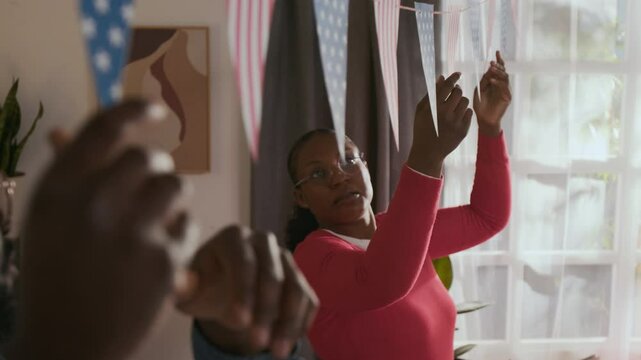 African American spouses both wearing glasses adjusting patriotic garland with flags after hanging it across room on independence day