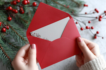 A red envelope held in delicate hands reveals a beautiful card nestled inside, set against a backdrop of festive pine branches and bright berries.