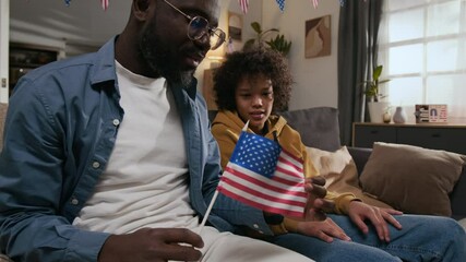 Gen alpha boy watching young dad waving small American flag and chatting with him while sitting on couch in living room - Powered by Adobe