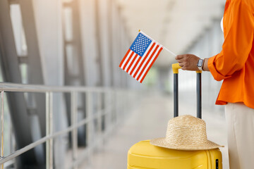Black female traveler holding American flag and standing by yellow suitcase with summer hat in...