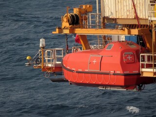 A vibrant orange lifeboat hangs suspended over a dark, choppy ocean. The vessel is attached to a yellow crane, ready for deployment. A sense of maritime safety and preparedness permeates the scene.