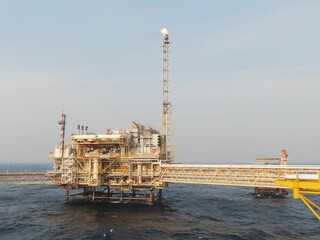 A majestic offshore oil platform stands tall against a serene, hazy sky, its flame a beacon in the vast ocean. The structure, complex and intricate, is a testament to human engineering prowess.
