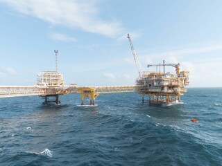 Offshore oil platform, yellow and white structures, on blue wavy ocean. A crane is visible. The background is a clear blue sky with some clouds. The atmosphere is industrial and powerful.