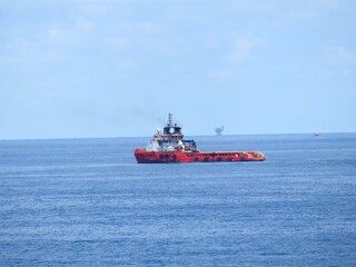 A red supply vessel navigates the vast ocean, a distant oil rig barely visible on the horizon under a pale sky. The calm sea reflects the peaceful atmosphere.