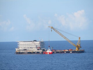 Offshore platform operation underway, a large crane lifts equipment. A smaller vessel assists nearby. The open ocean under a mostly clear sky provides a dramatic backdrop to this industrial scene.