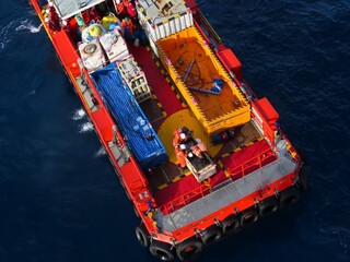 Workers in orange and red safety gear meticulously manage cargo on a vibrant red and yellow vessel, floating on a deep blue ocean. The scene depicts offshore industrial operations.