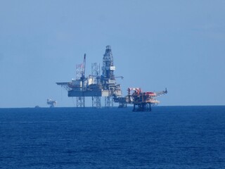 Oceanic Oil Platforms Under Vast Azure Sky AStunning View Of Offshore Drilling Rigs Against The Deep Blue Sea.Tranquil And Imposing Presence