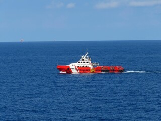 Red and white vessel navigating the deep blue ocean, under a clear sky. A distant platform is barely visible on the horizon. The ship cuts through the water, leaving a small wake.